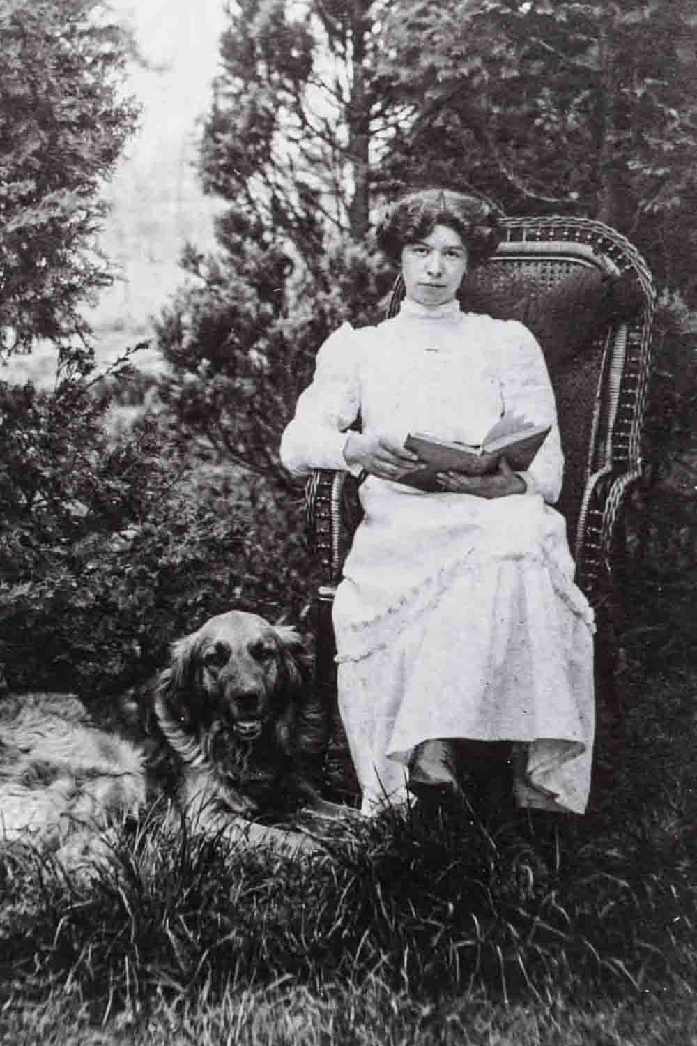 Een portret van dichteres Margot Vos. Zij zit buiten in een grote stoel met een boek in haar handen. Ze kijkt in de camera. In het gras naast haar ligt een grote hond.